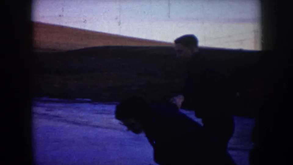 1953 North Dakota home-movie frame showing two adults in dark winter clothing skating on a frozen pond.