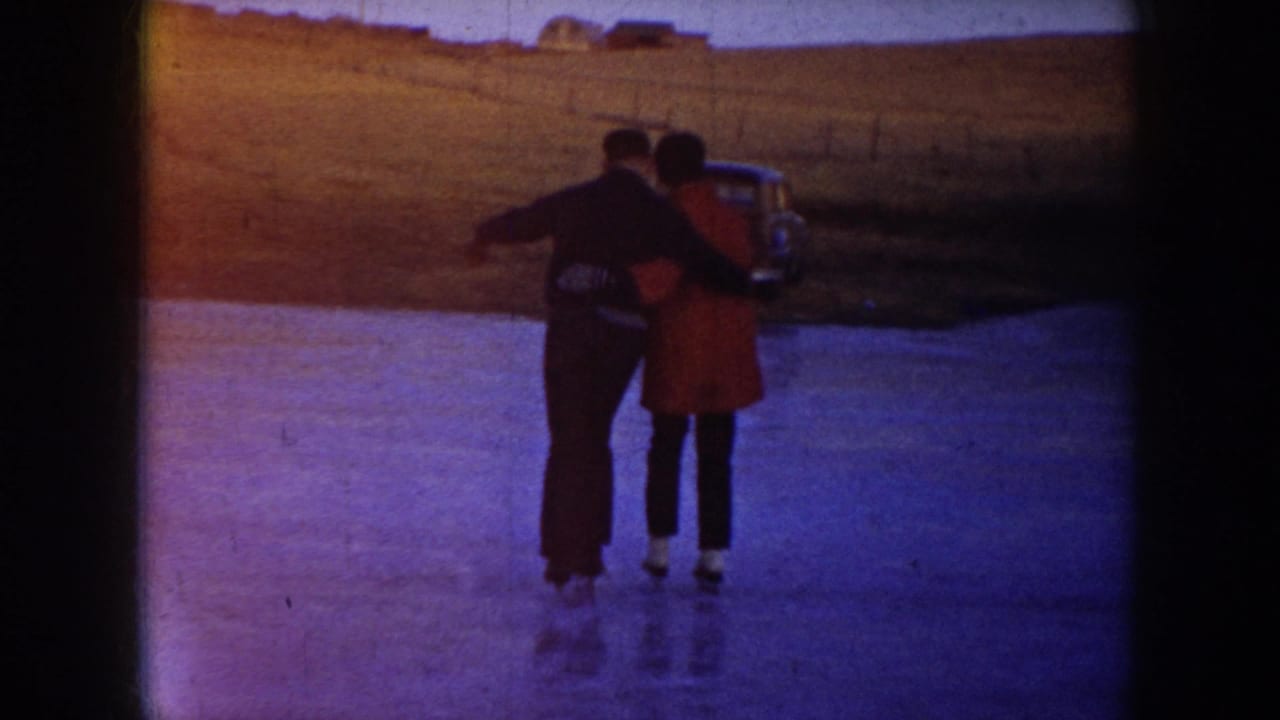 Two skaters in winter clothing hold hands on a frozen pond in North Dakota, with a mid-century car near open fields behind them.