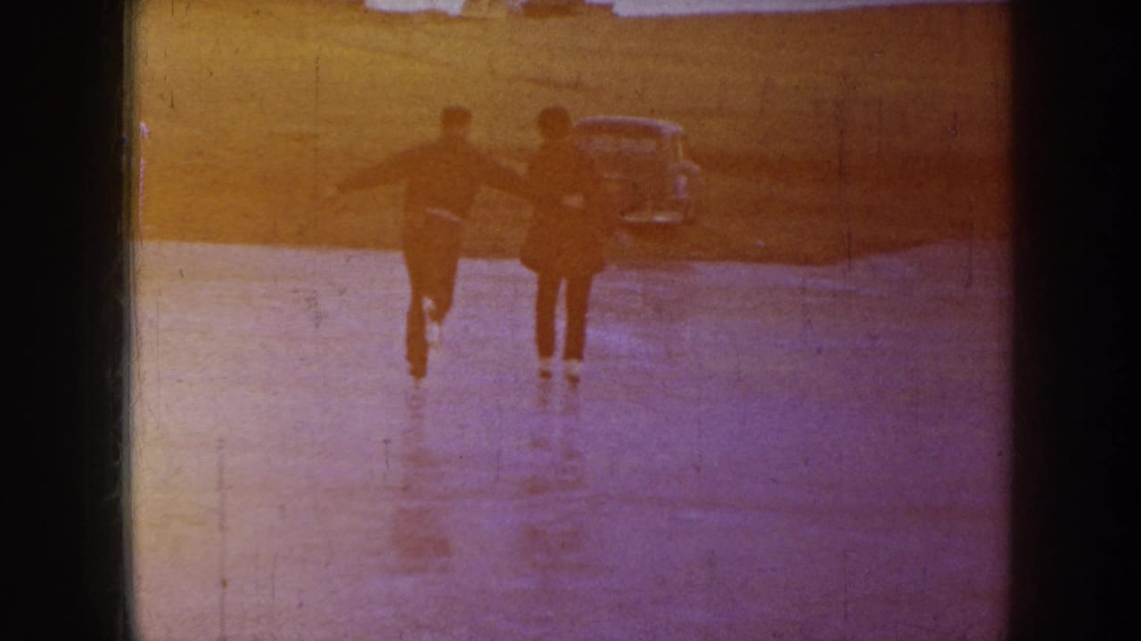 Two people skate away across a sunlit frozen pond with a car behind them.