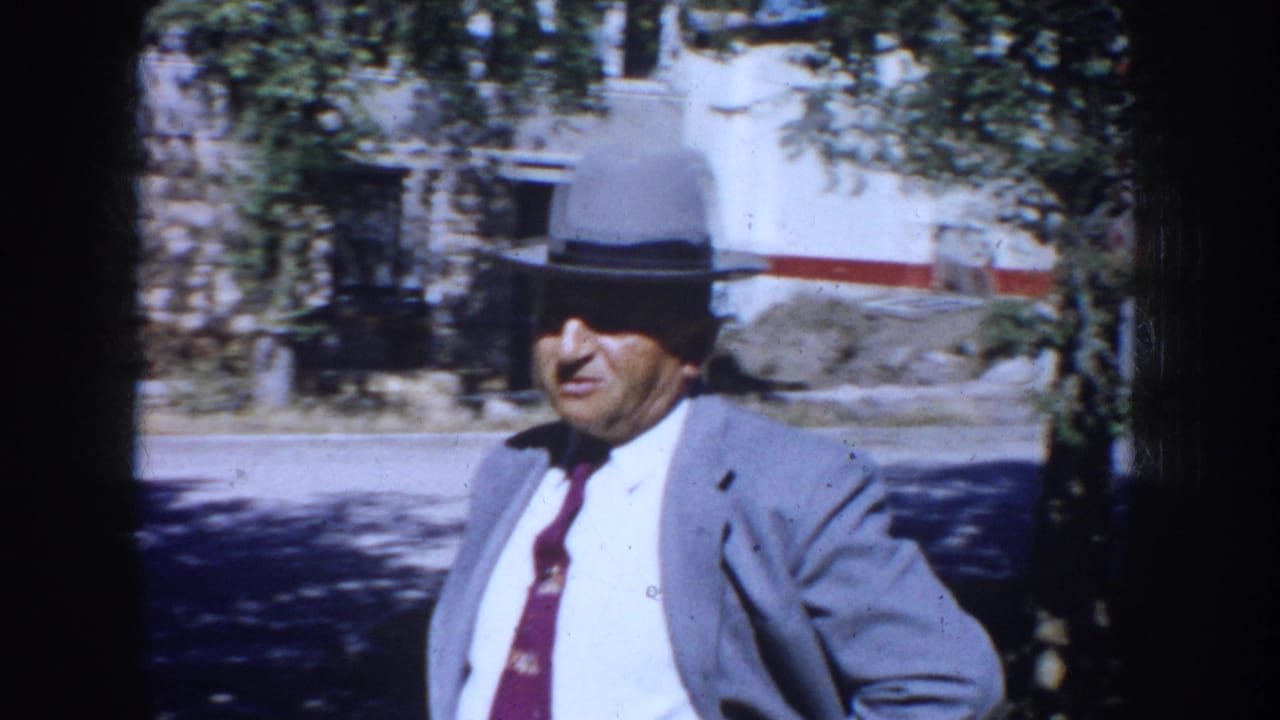 Older man in a gray fedora and suit walking outdoors near trees and a building.
