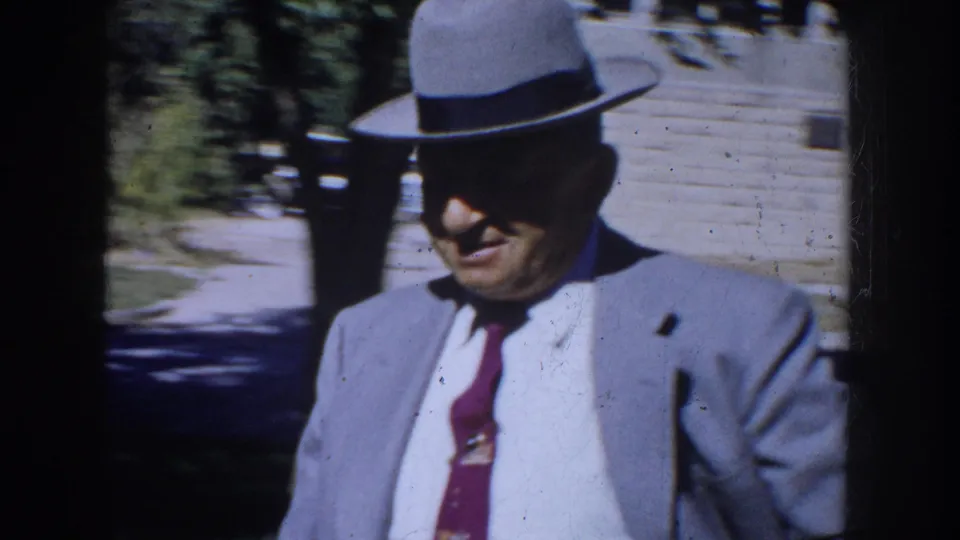 Closer 1953 South Dakota home-movie view of an older man in a gray fedora, suit jacket, white shirt, and red tie walking outdoors.