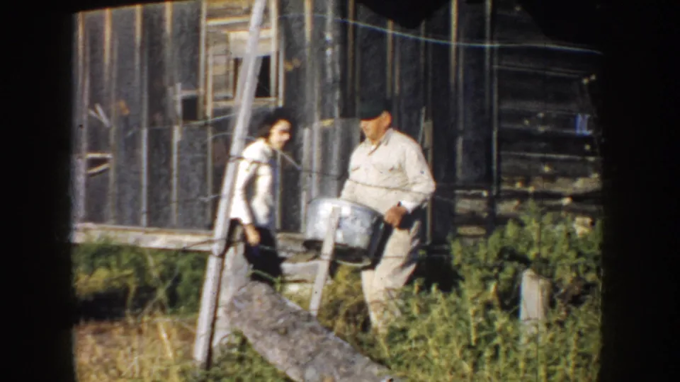 A woman and a man stand by the weathered barn, and the man holds a metal pail during the rural horse-handling moment.