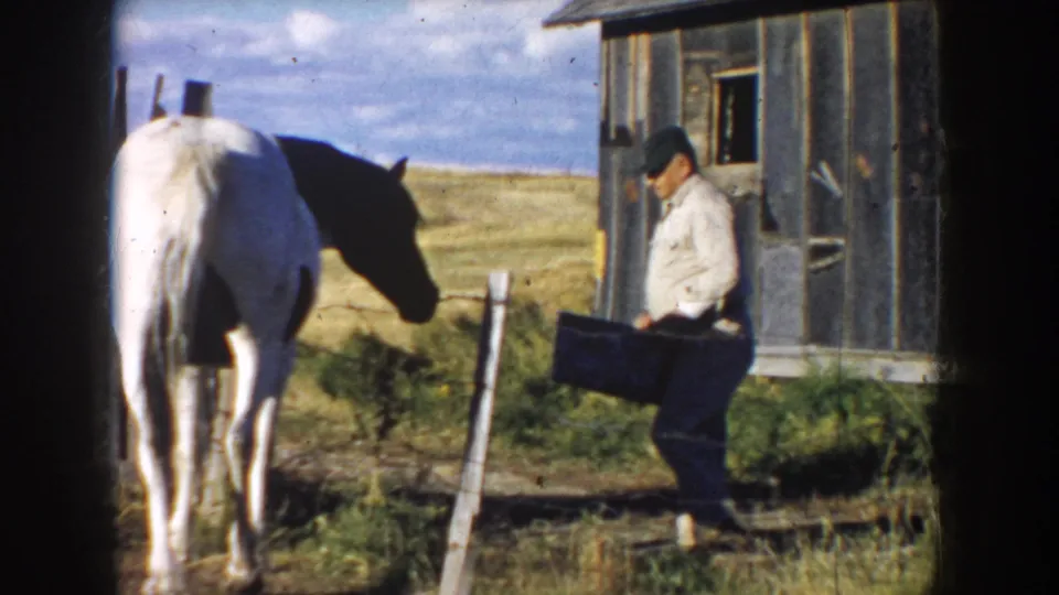 1953 South Dakota 8mm frame showing a farmer with a bucket beside a white horse near a weathered wooden barn and open pasture.