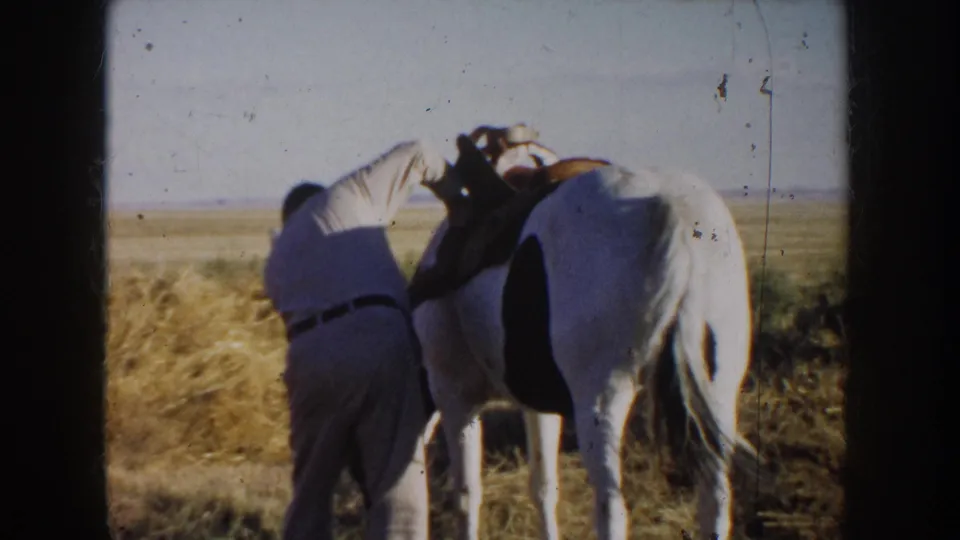 1953 South Dakota home-movie frame showing a man beside a white and dark-marked horse while preparing tack near hay.