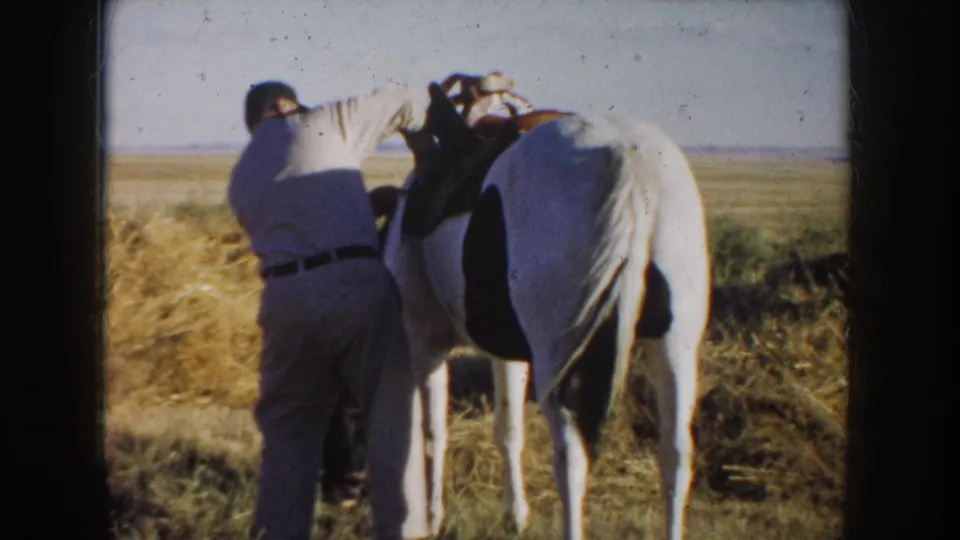 1953 South Dakota home-movie frame showing a man in a light shirt and dark cap working beside a saddled white horse in dry grass.