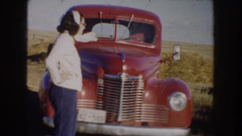 1953 South Dakota home-movie frame with a young woman in a light sweater beside a red pickup in prairie grass.