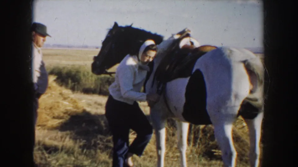 1953 South Dakota frame showing a woman working at the saddle of a pinto horse while a man in a cap stands nearby.