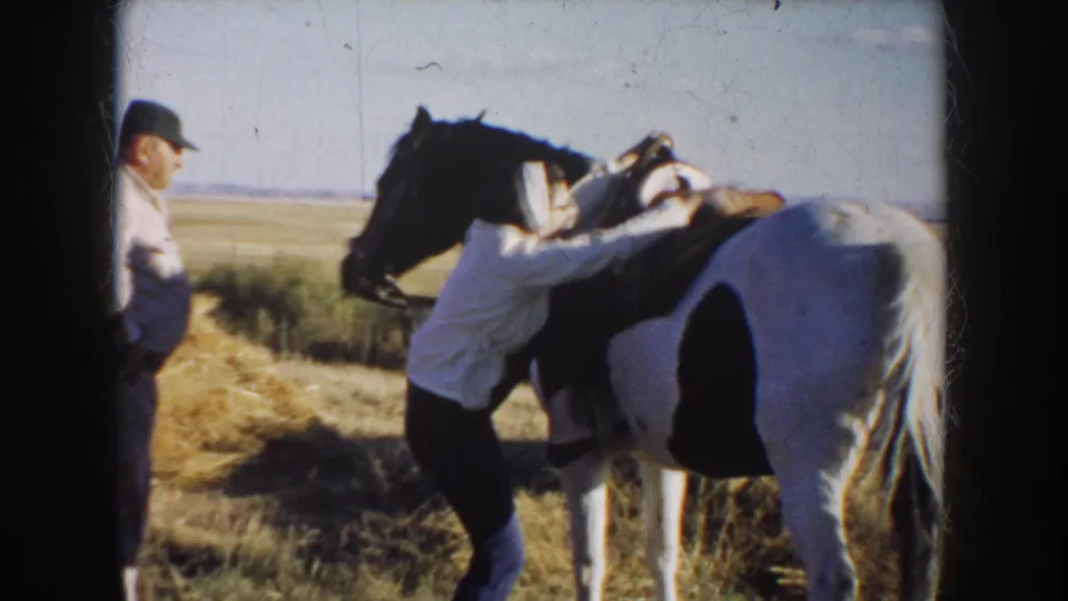 1953 South Dakota home-movie frame with a woman leaning over a saddled pinto horse in an open field as a man watches.