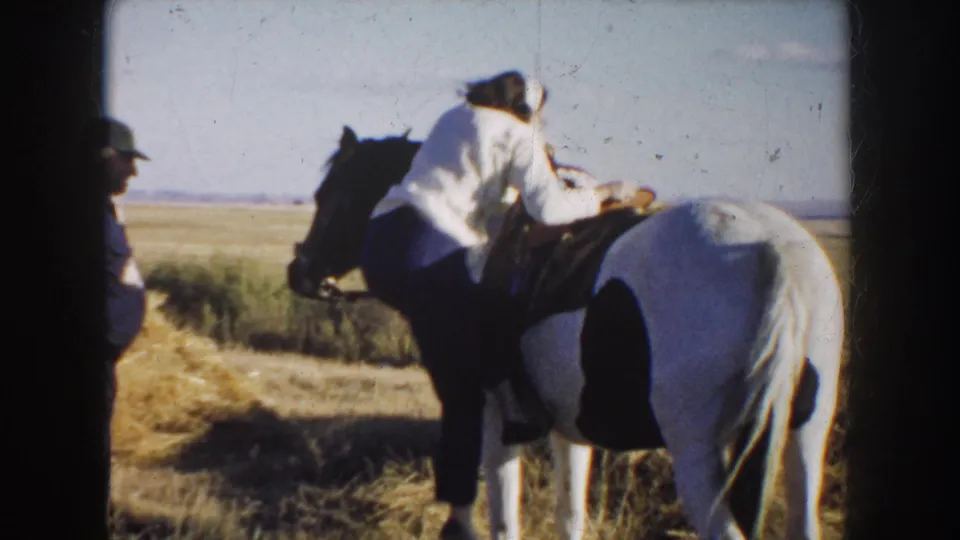 In South Dakota 1953 home-movie footage, a woman in a light blouse climbs onto a saddled pinto horse in an open field.