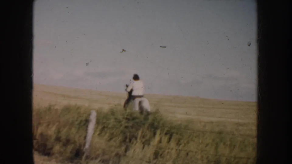 Distant rider and pinto horse on South Dakota prairie, with foreground fence posts and aged 8mm film texture.