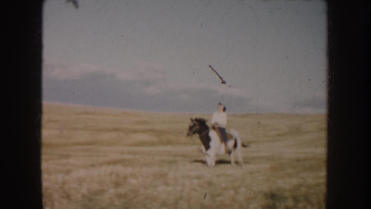 Soft 8mm frame from South Dakota showing a rider on a pinto horse in golden pasture, with open land beyond.
