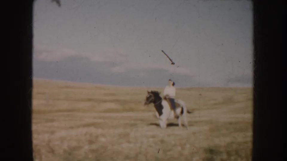 Soft 8mm frame from South Dakota showing a rider on a pinto horse in golden pasture, with open land beyond.