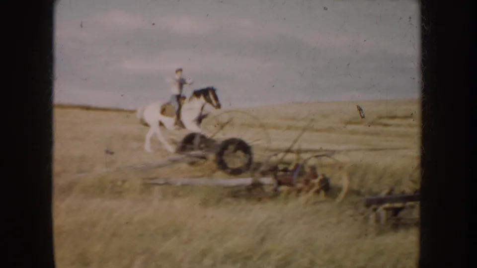 A man rides a pinto horse past scattered farm implements in a South Dakota field, seen in soft 8mm color.