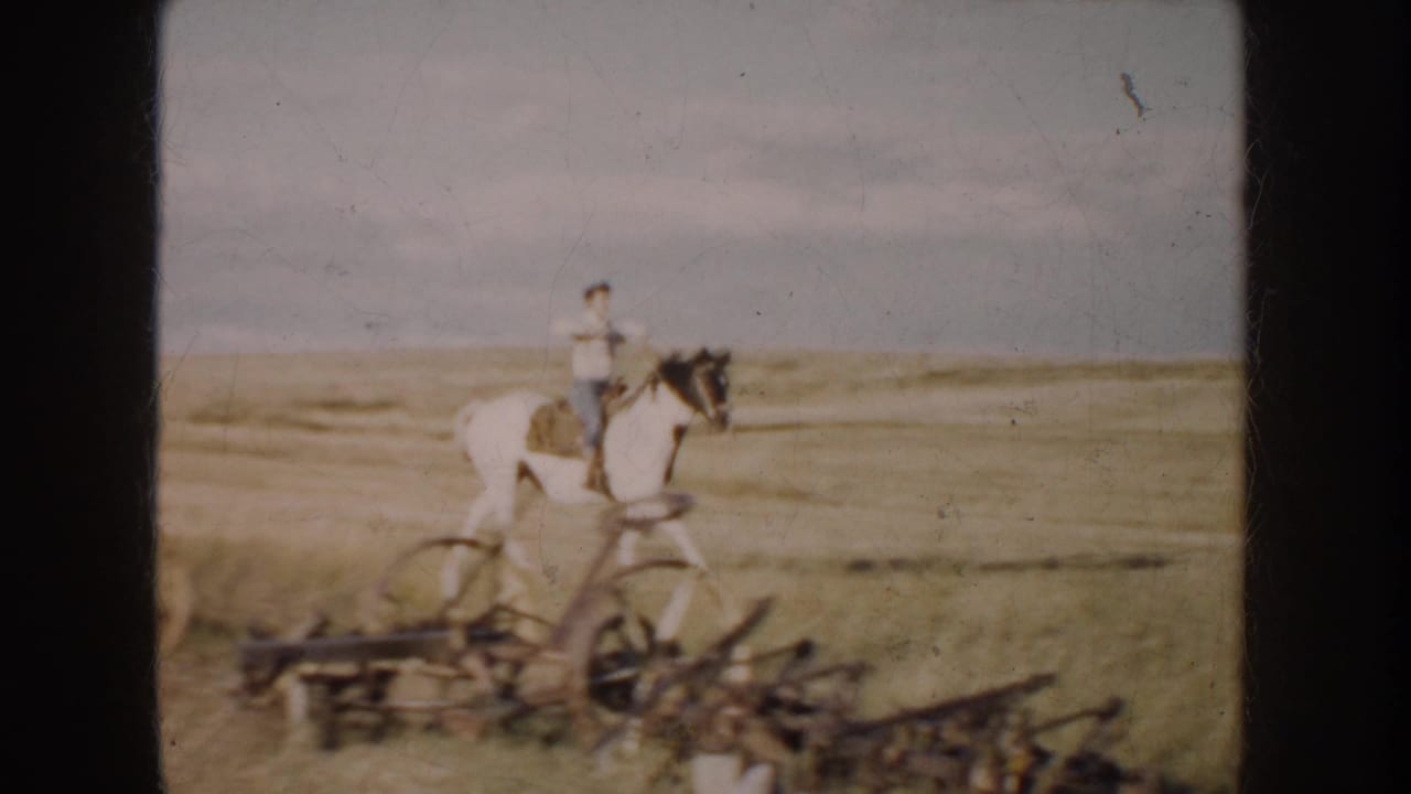 1950s rural frame of a man riding a black-and-white horse beside low farm machinery on South Dakota grassland.