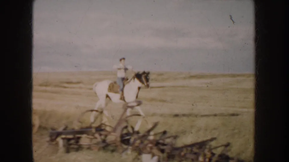 1950s rural frame of a man riding a black-and-white horse beside low farm machinery on South Dakota grassland.