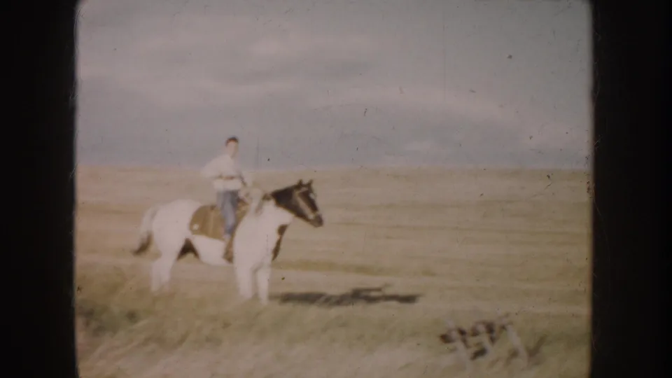 A man on a pinto horse appears in open South Dakota grassland, with small farm equipment visible near the frame edge.
