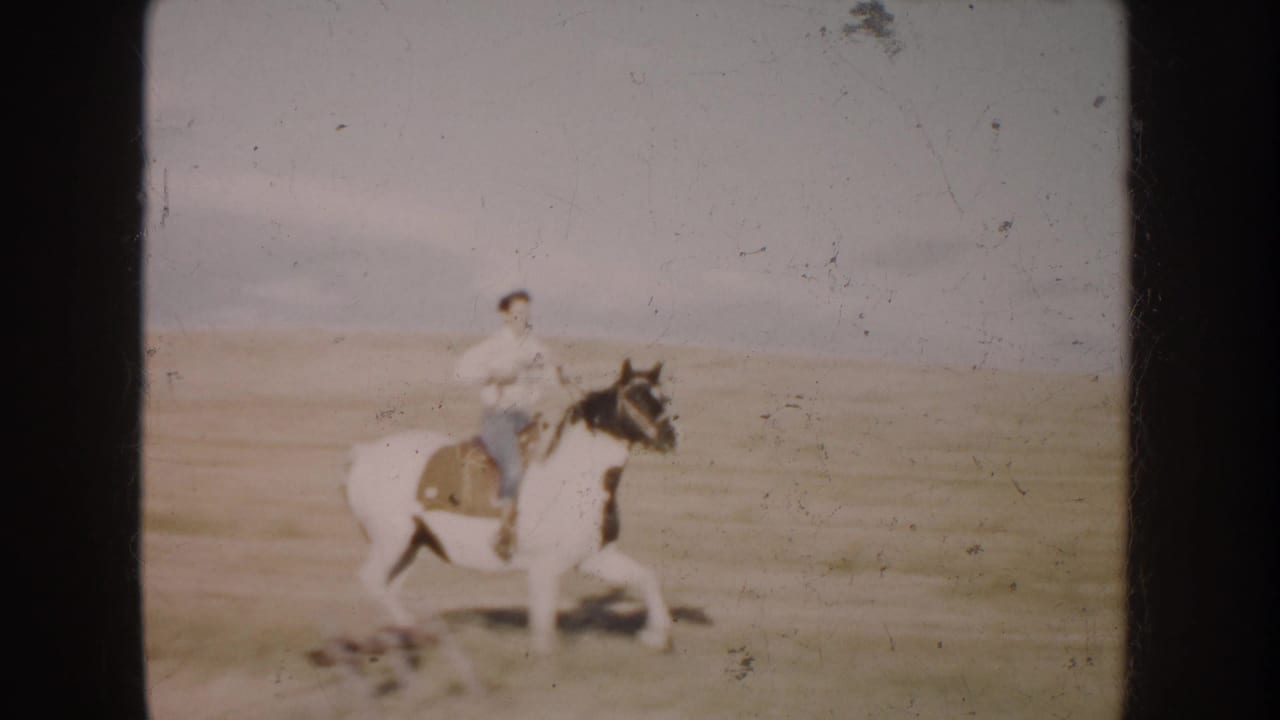 In South Dakota home-movie footage from 1953, a young rider guides a pinto horse across a grassy field under a pale sky.