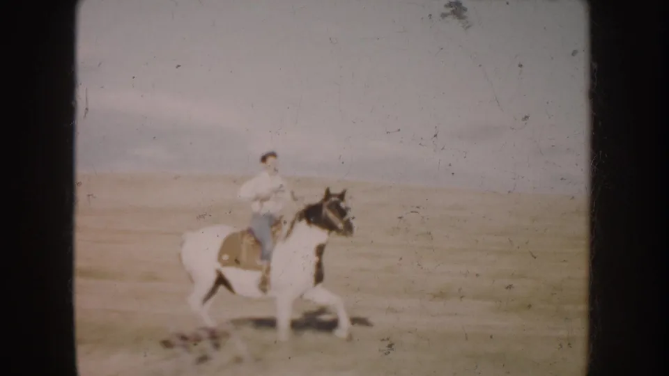 In South Dakota home-movie footage from 1953, a young rider guides a pinto horse across a grassy field under a pale sky.