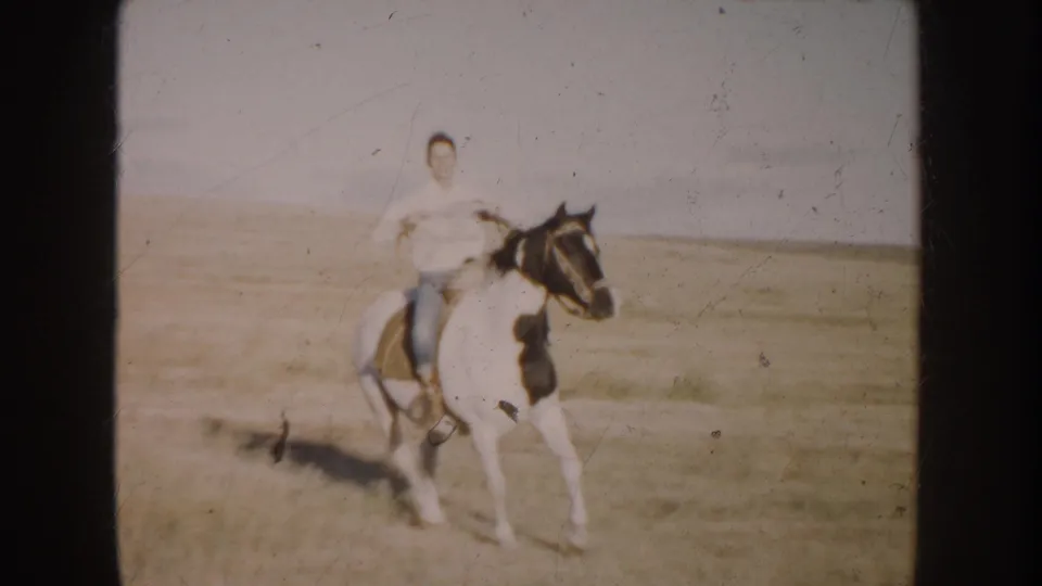 The rider and black-and-white pinto horse appear nearer the camera, with saddle and reins visible against the South Dakota grassland.