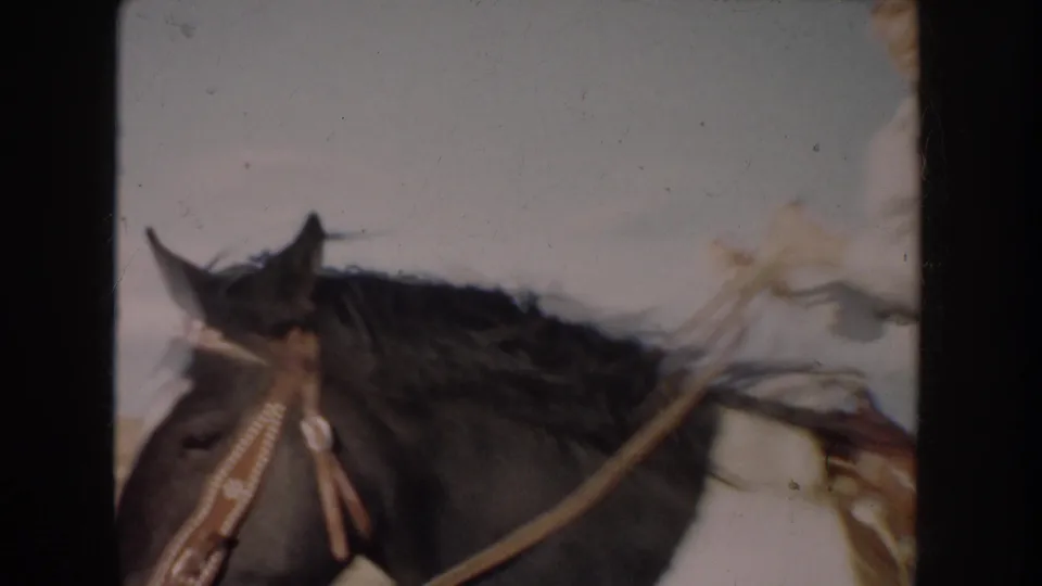 A close handheld frame crops in on the pinto horse's head, bridle, reins, saddle, and the rider's jeans as they pass nearby.