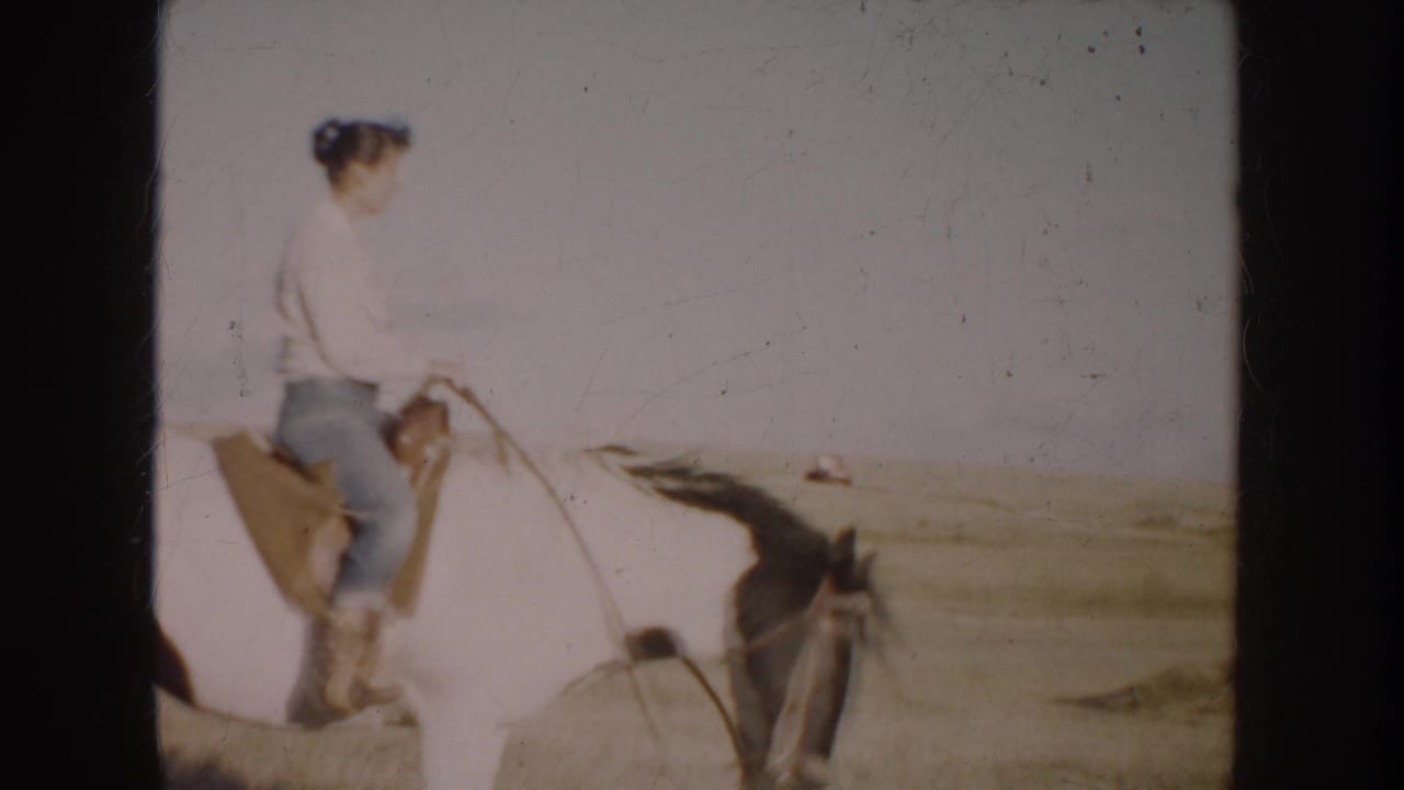 1953 South Dakota 8mm frame of a woman in casual clothes riding a white horse across prairie, with a distant red pickup in the background.