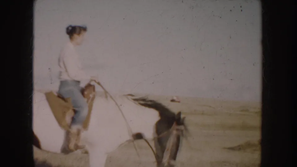1953 South Dakota 8mm frame of a woman in casual clothes riding a white horse across prairie, with a distant red pickup in the background.