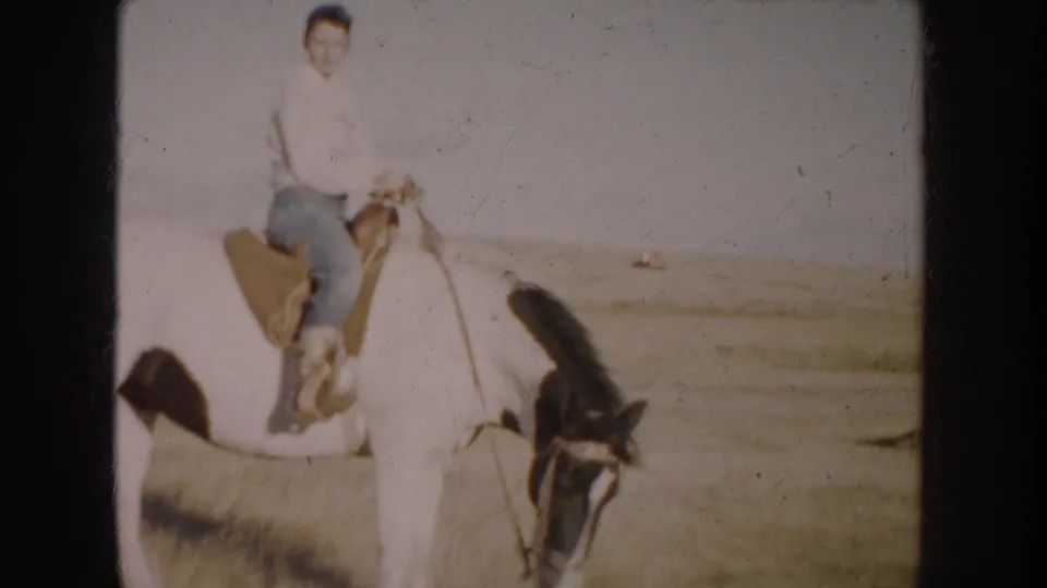 1953 South Dakota home-movie frame showing a woman seated on a white horse, facing the camera across sunlit prairie land.
