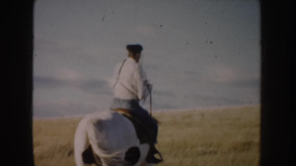 Rear view of a woman riding a dark brown and white horse through South Dakota grassland in 1953 home-movie footage.