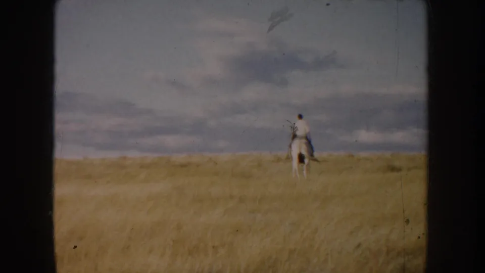 1953 South Dakota 8mm frame showing a lone rider on a white horse crossing open golden prairie under layered clouds.