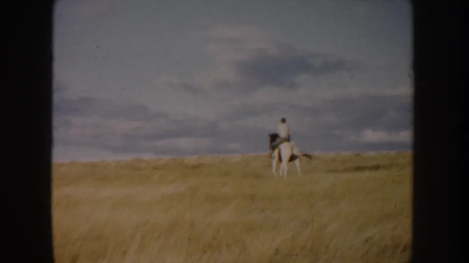 Distant rider on a white horse continues across a South Dakota prairie field in faded 1950s 8mm color.