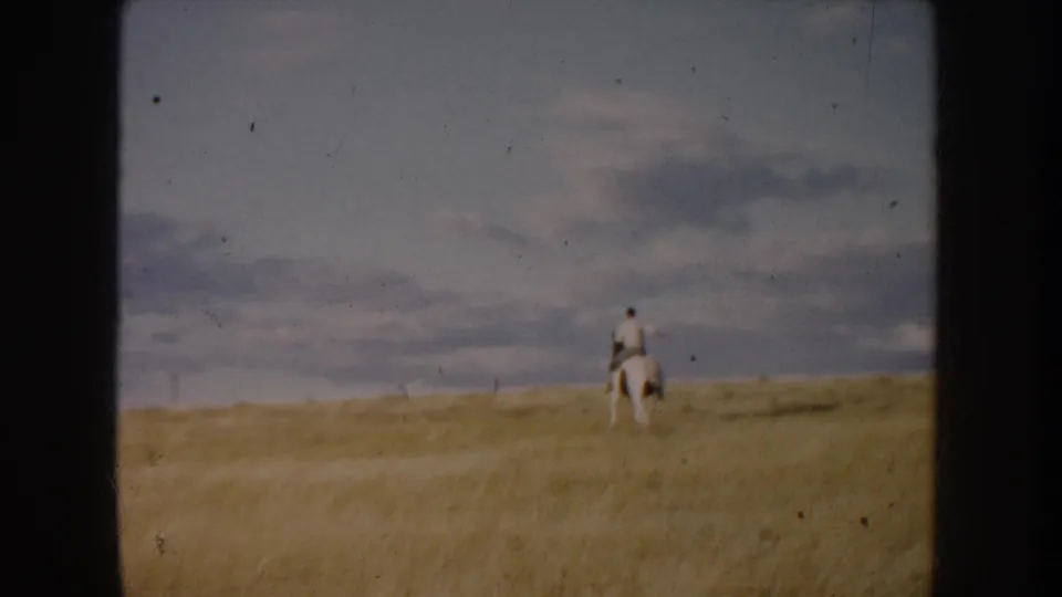 Home-movie frame from South Dakota in 1953 showing the rider from behind as the white horse moves through tall prairie grass.