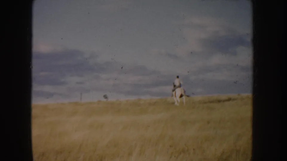 A lone horseback rider appears near the horizon on open South Dakota prairie, framed by golden grass and a cloudy 1950s 8mm sky.
