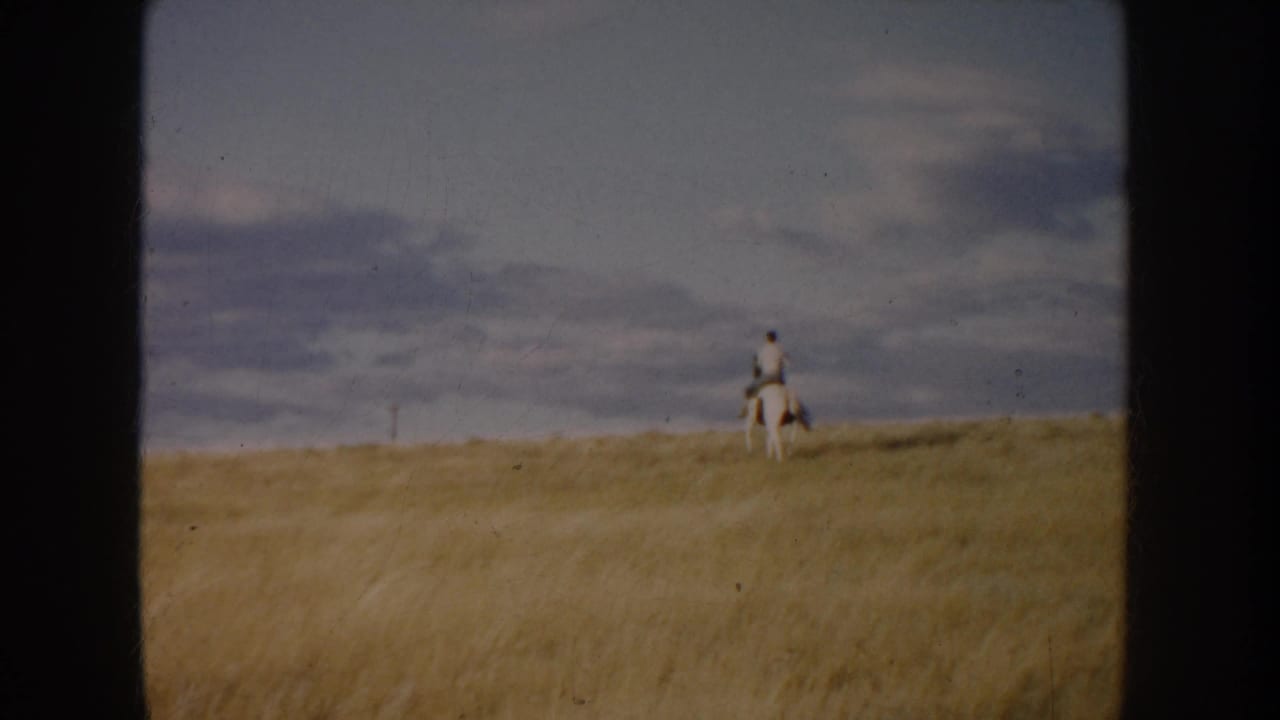 In 1953 South Dakota footage, a distant rider on a white horse moves along a golden grassy rise beneath a clouded sky.