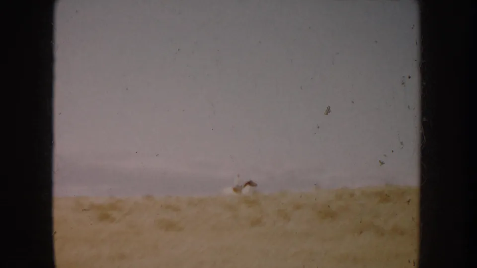 South Dakota home-movie still showing a small distant horse and rider low against a dusty field and gray sky.