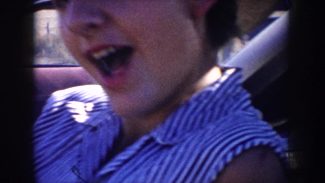 In 1953 South Dakota home-movie footage, a woman in a green hat and striped dress reacts playfully from inside a car.