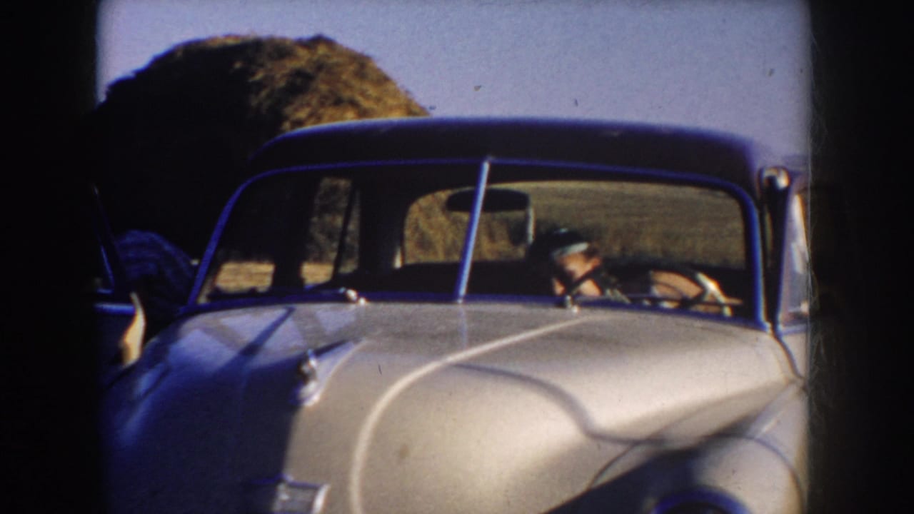 The home movie frames a pale classic car from the front, with a hat-wearing man seen through the windshield in rural South Dakota.