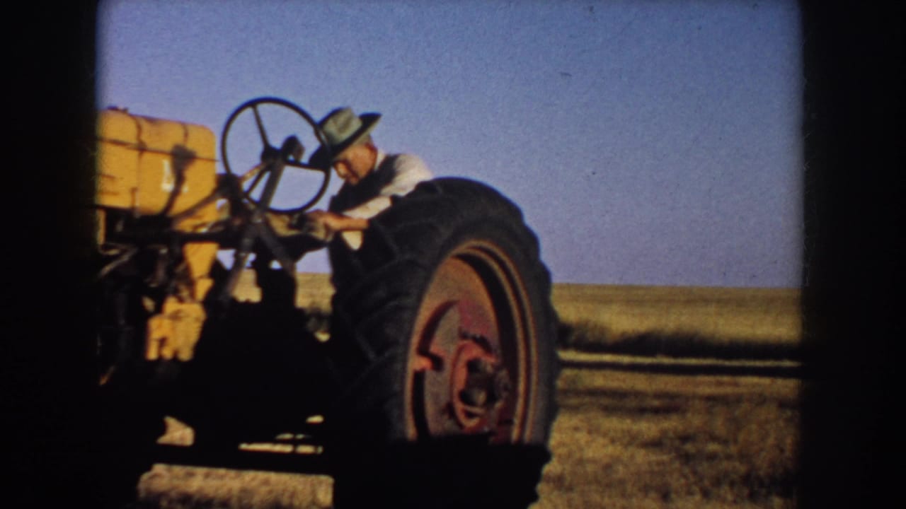 A man in a hat works beside or starts a yellow tractor in a South Dakota field, with open sky and prairie behind him.