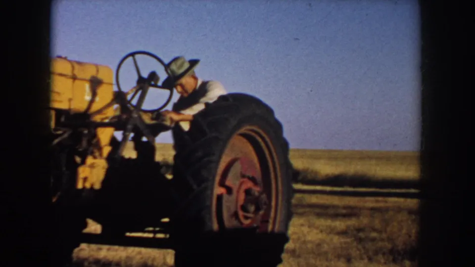 A man in a hat works beside or starts a yellow tractor in a South Dakota field, with open sky and prairie behind him.