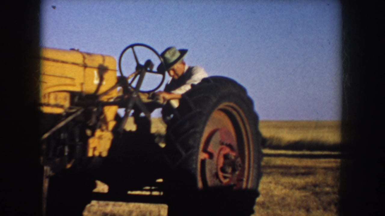 1953 South Dakota home-movie frame showing a farmer in a hat near the steering wheel of a yellow tractor in open farmland.