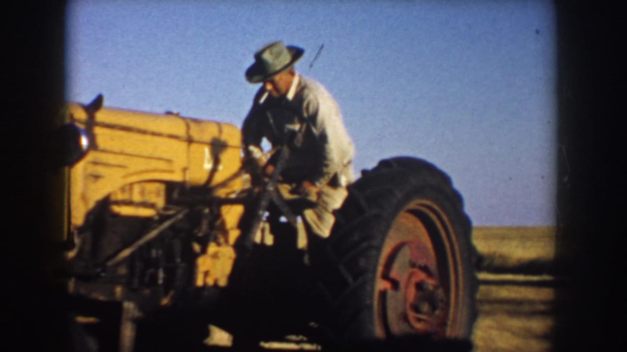 Farmer with a cigarette stands on a yellow tractor under a blue sky.