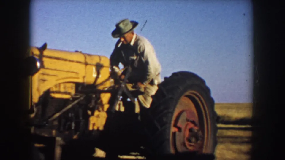 A farmer in a wide-brimmed hat and light work clothes appears to stand on or climb onto a yellow tractor in South Dakota, 1953.