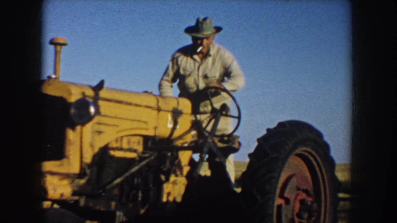 South Dakota 1953 frame showing a farmer standing beside the steering wheel and hood of a yellow tractor in a broad rural field.