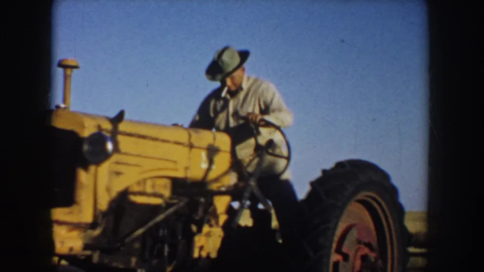 A 1953 South Dakota home-movie frame shows the farmer looking down at the tractor controls while seated or climbing onto the yellow tractor.