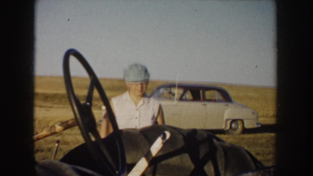 Young person in a cap near farm equipment, with a pale sedan in the background.