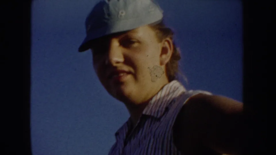 Close home-movie portrait of a young woman or teen in a blue cap and striped sleeveless shirt against the South Dakota sky.