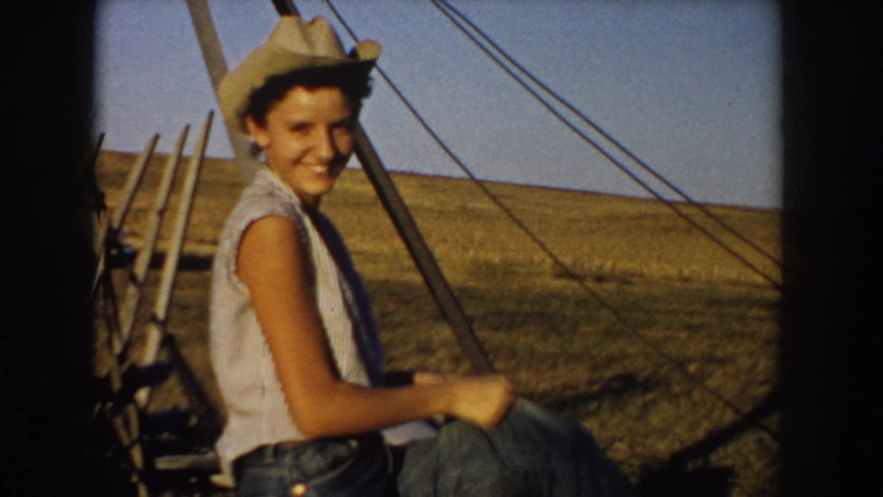 Smiling teen in a cowboy hat sits near hay and farm equipment in a 1953 South Dakota rural field scene.