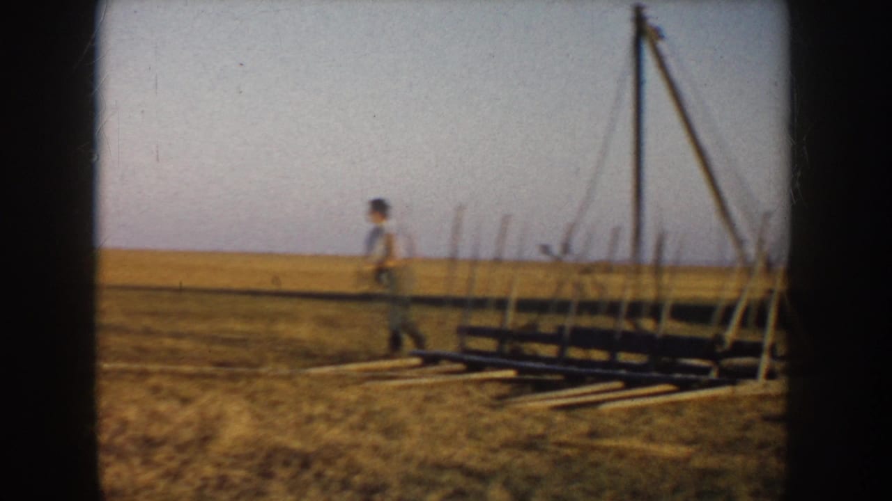 A man in a rural South Dakota field near a wooden hay-handling frame, seen in soft 8mm color.