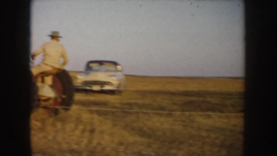 1953 South Dakota home-movie frame showing a tractor driver near a parked period car on an open prairie field.
