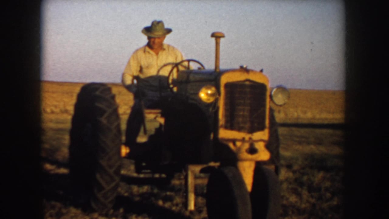 Man wearing a hat drives a yellow tractor through an open South Dakota prairie field in 1953 home-movie footage.