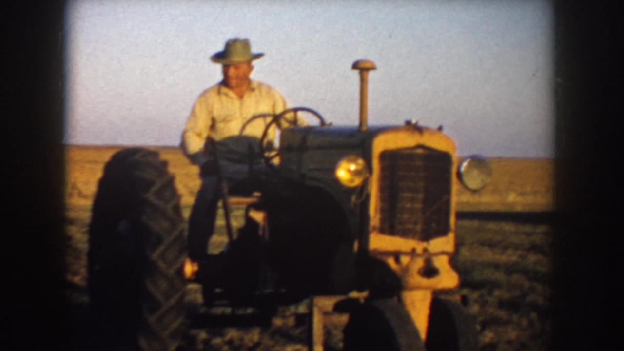 In 1953 South Dakota home-movie footage, a farmer in a light shirt and wide-brimmed hat drives a vintage tractor through a sunlit field.